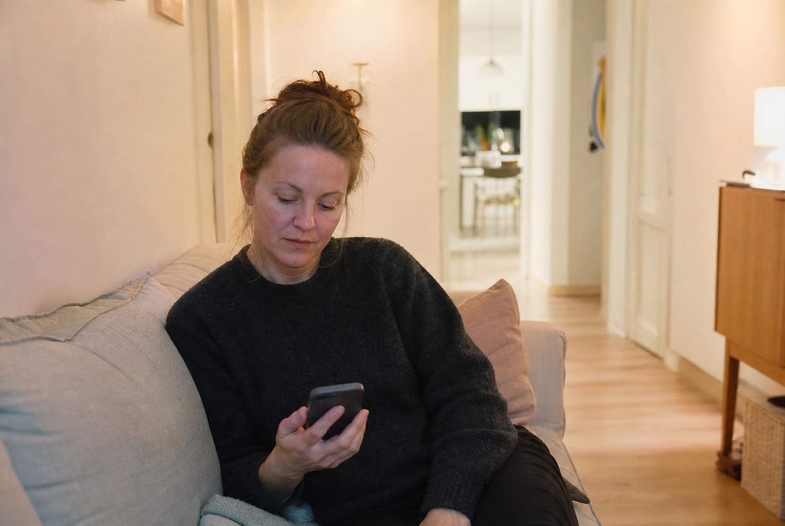 En kvinna sitter i soffan i ett ljust svenskt vardagsrum på kvällen och tittar ner i sin mobil.